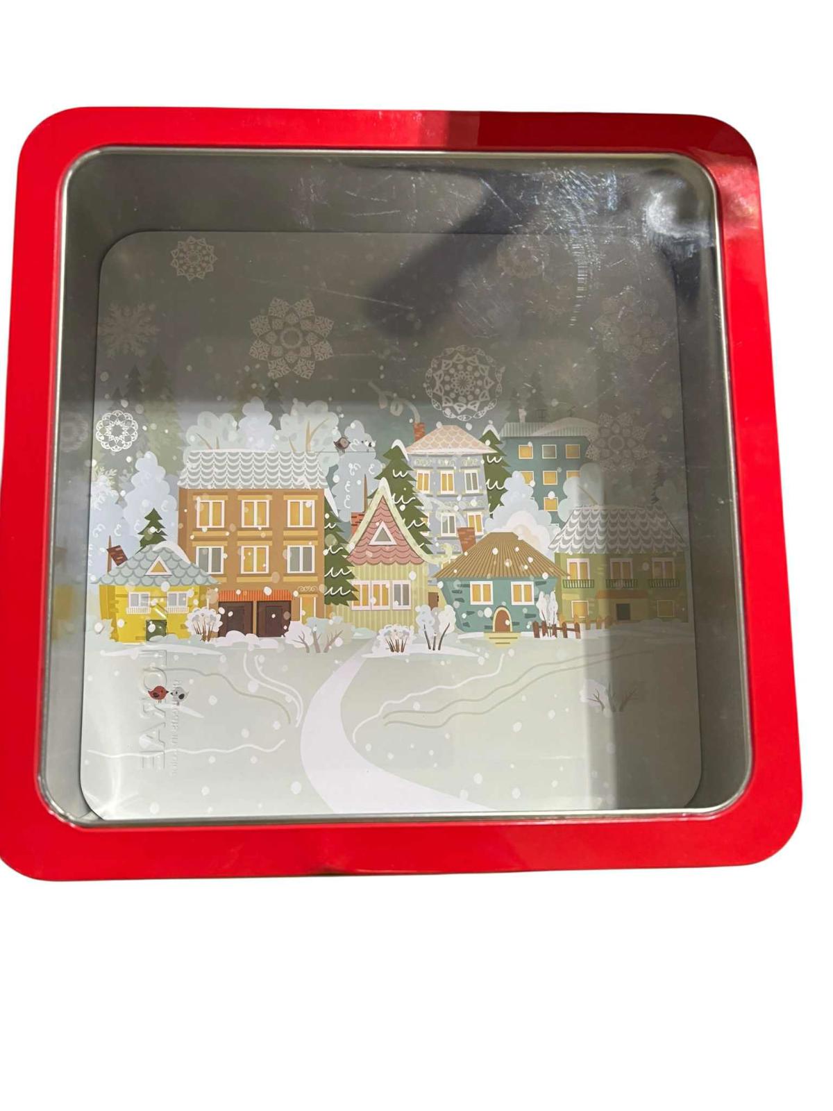 Holiday-themed red tin container featuring a snowy village scene with colorful houses.
