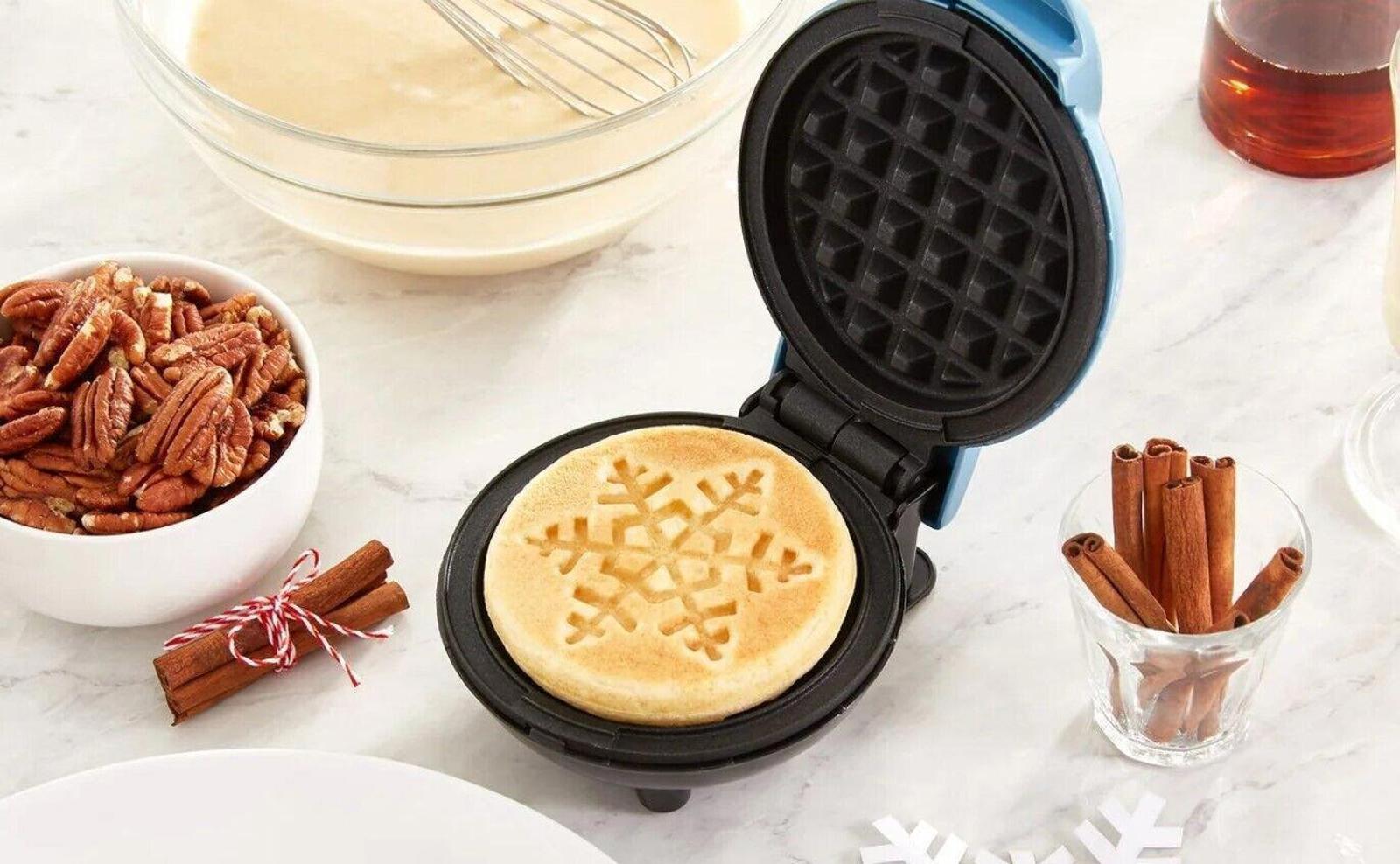 Waffle maker with snowflake design waffle, surrounded by pecans and cinnamon sticks.