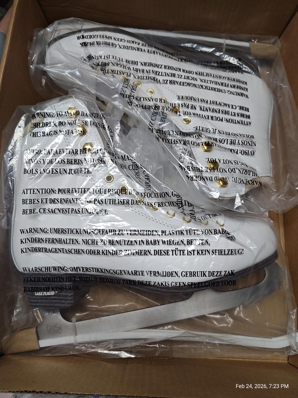 White ice skates with gold accents in a box, featuring safety warnings on the packaging.
